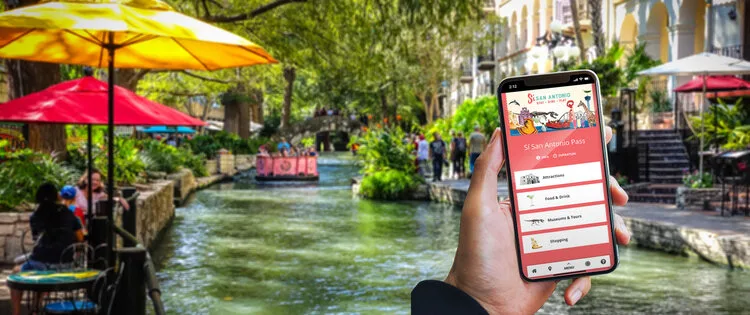Hand holding a smartphone with the Visit San Antonio app above the River Walk and its colorful umbrellas