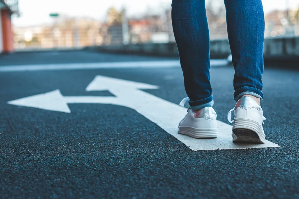 Person in jeans and white sneakers walking at a fork of road arrows pointing left and right