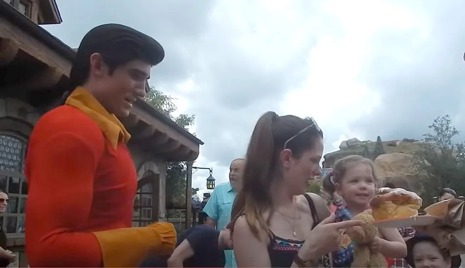 Gaston character presenting a giant cinnamon bun to a toddler held by her mother at a Disney park