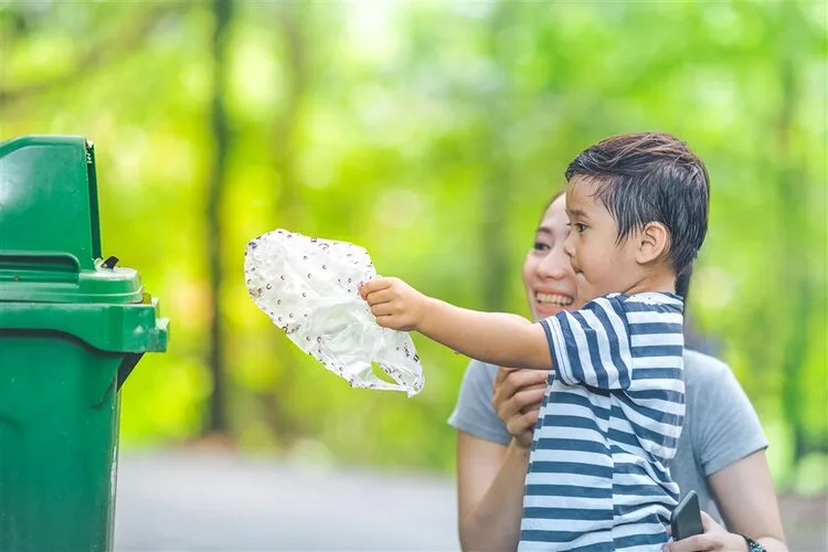 Child tossing a plastic bottle into a green trash bin while held by a parent in a park