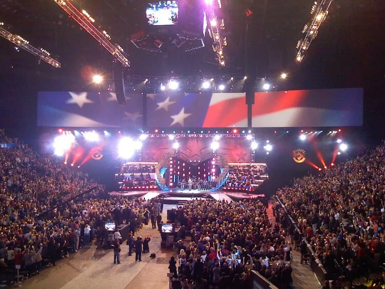 Packed arena facing a stage with an American flag video wall at the Oklahoma birthday celebration