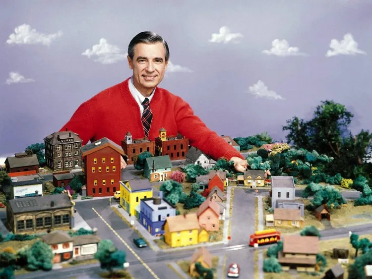 Mister Rogers' Neighborhood host in a red cardigan with the show's model neighborhood of houses and trolley