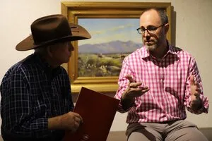 Man in a cowboy hat and man in a checked shirt in conversation before a landscape painting