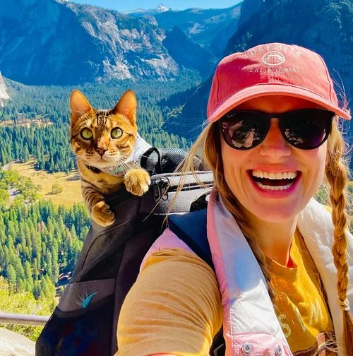 Smiling hiker taking a selfie with her cat riding in a backpack above a mountain valley