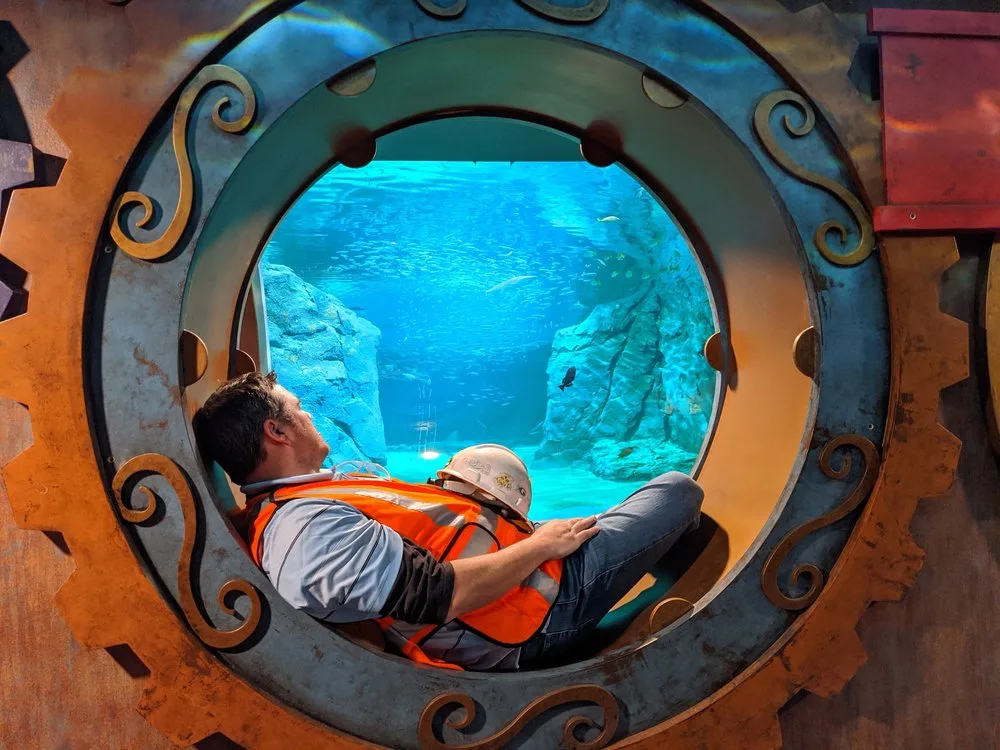 Construction worker in a hard hat resting in a gear-framed porthole window overlooking an aquarium tank