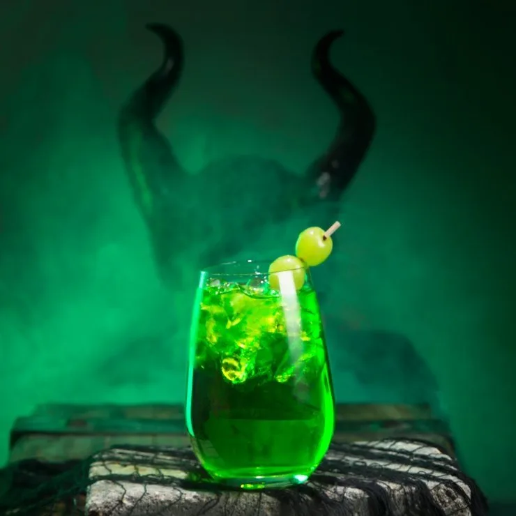 Glowing green Maleficent-themed melon whiskey sour against a smoky backdrop with horned silhouette