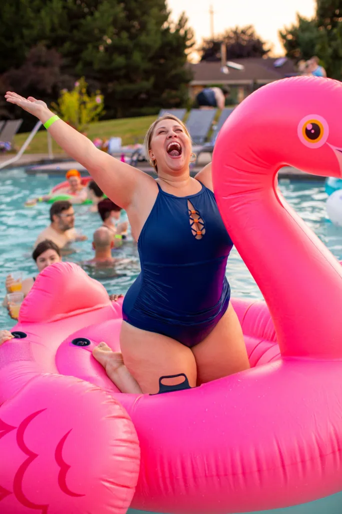 Joyful woman in a swimsuit striking a pose on a giant pink flamingo float at a pool party