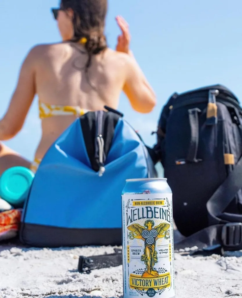 Can of WellBeing Victory Wheat non-alcoholic brew in the sand beside a beachgoer and backpacks