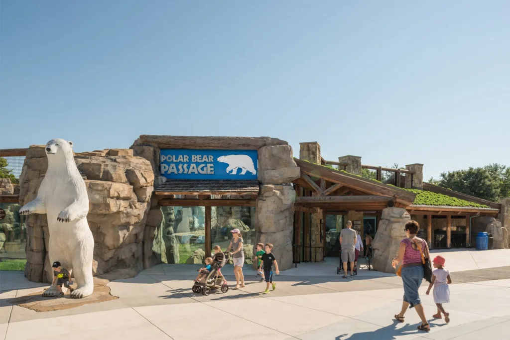 Families walking past a polar bear statue at the Polar Bear Passage entrance at Kansas City Zoo, topped by a green roof