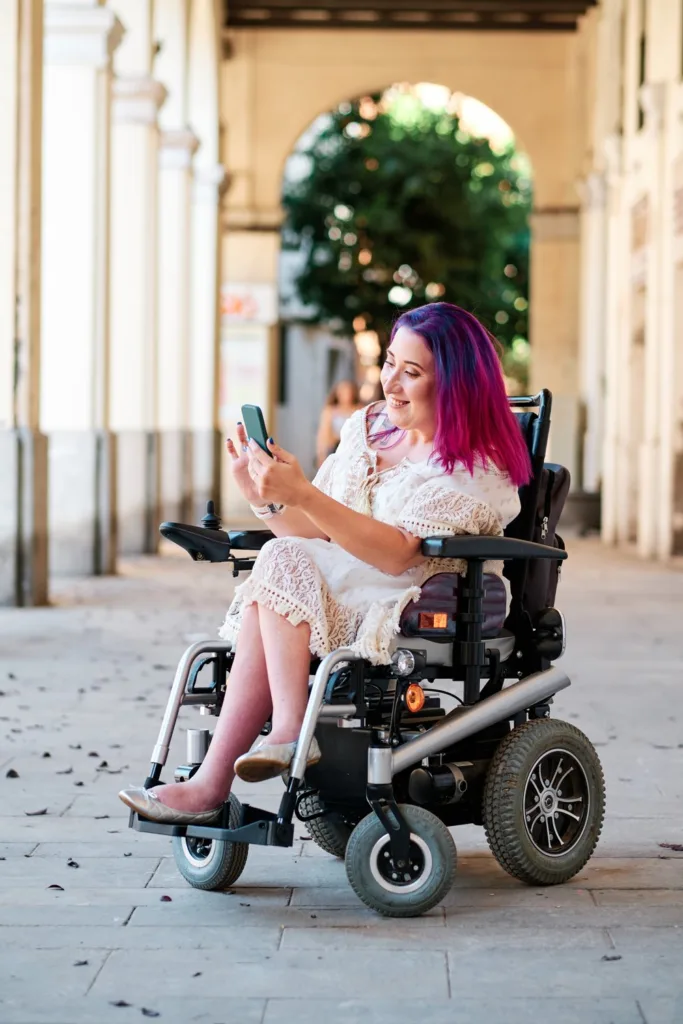 Woman with pink hair in a power wheelchair smiling at her phone in an arched colonnade