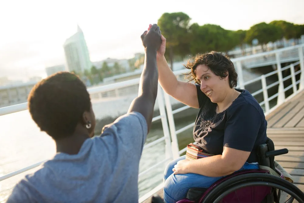 Woman in a wheelchair high-fiving a friend on a waterfront boardwalk