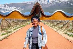 Smiling traveler on a vineyard path in front of a winery with a wavy roof and mountains behind