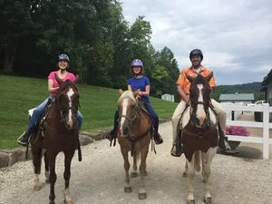 Three riders on horseback posing on a gravel path beside a white fence and green lawn
