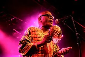 Performer in a patterned African print shirt singing on stage under pink lights