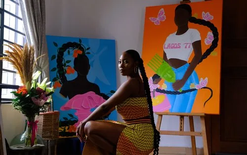 Woman with long braids seated in an art studio beside bold, colorful contemporary portraits