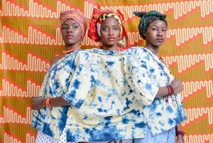 Three women in tie-dyed garments and head wraps posing before a vibrant patterned backdrop