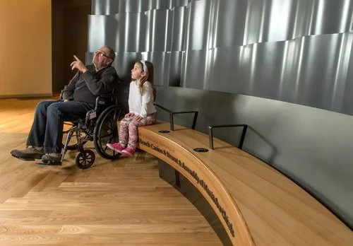 Man in a wheelchair and a child beside a curved wood bench with grab rails and an audio keypad jack