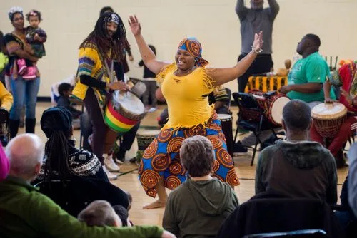 Dancer in a yellow top and African print skirt performing with drummers for a seated audience