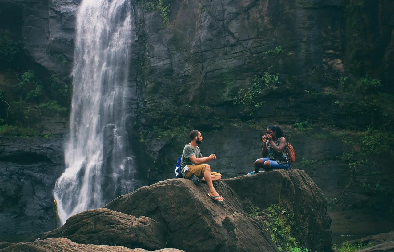 Two hikers resting on a boulder beside a jungle waterfall