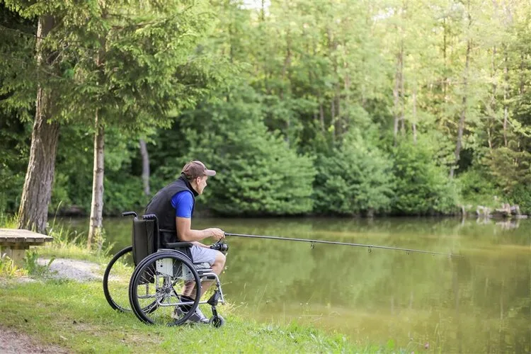 Man in a wheelchair fishing from the grassy bank of a wooded pond