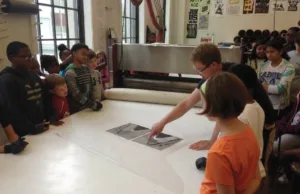 Young students gather around a large machine and listen to a design lecture