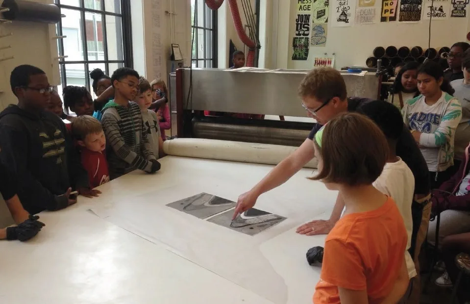 Young students gather around a large machine and listen to a design lecture