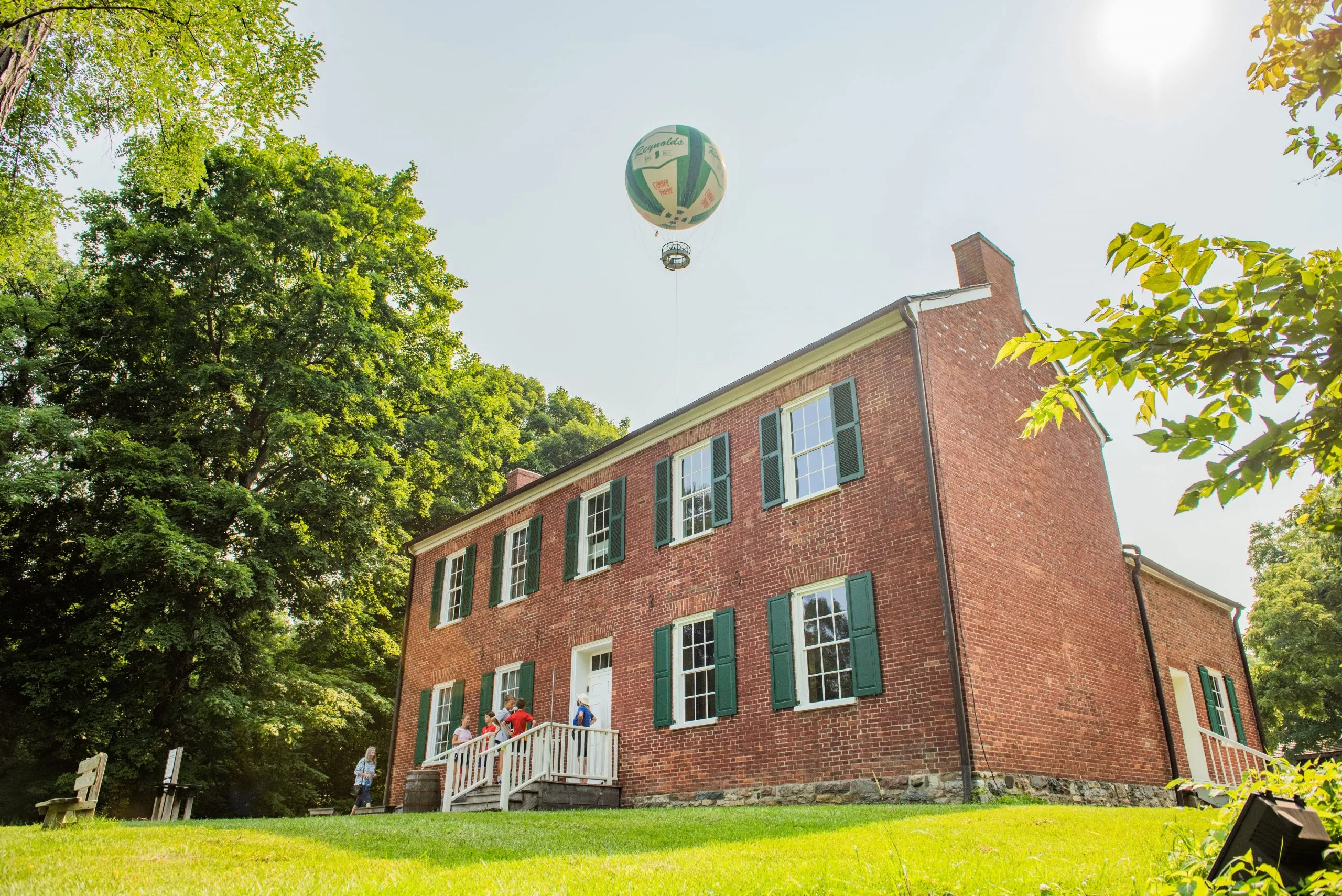 Historic red-brick house with green shutters at Conner Prairie, with a tethered balloon in the sky above