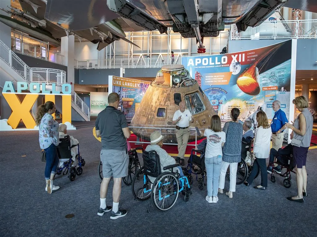 Visitors, some in wheelchairs, gathered around the Apollo XII command module while a guide talks about the mission