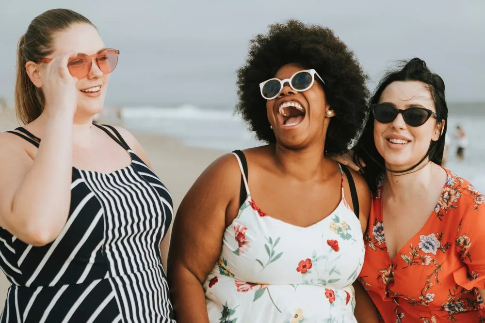 Three smiling women in sunglasses laughing together on a beach