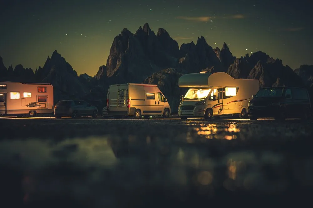 Camper vans parked beneath jagged mountains at night, their windows glowing, reflected in still water