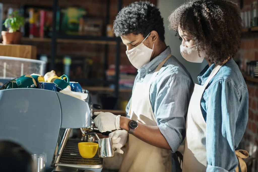 Masked barista in gloves training a coworker on an espresso machine in a cafe