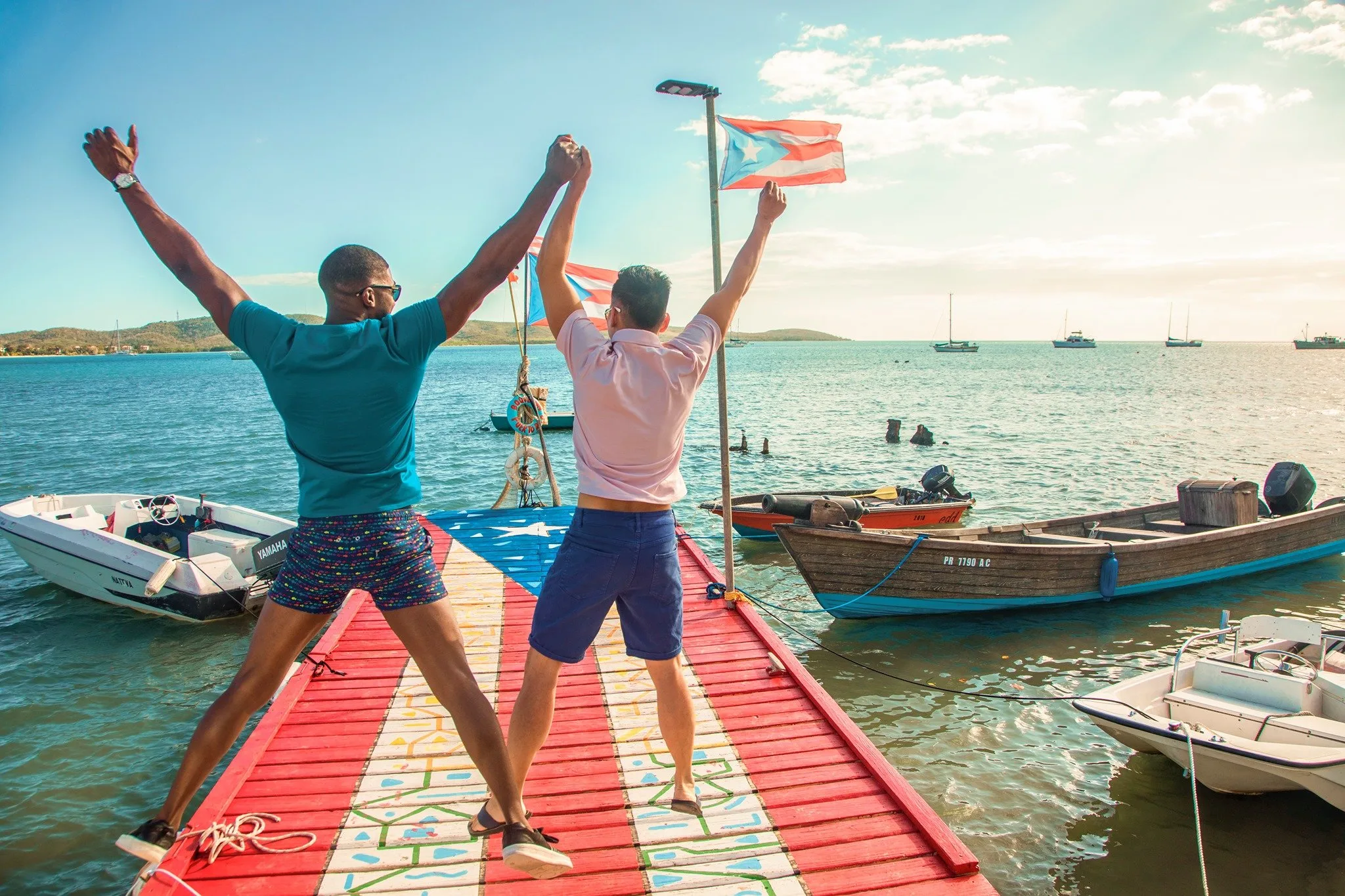 Two men cheering with arms raised on a dock painted like the Puerto Rican flag beside moored boats