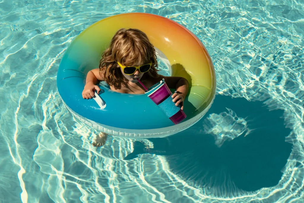 Child in sunglasses floating on a rainbow inner tube in a sparkling pool, sipping a drink