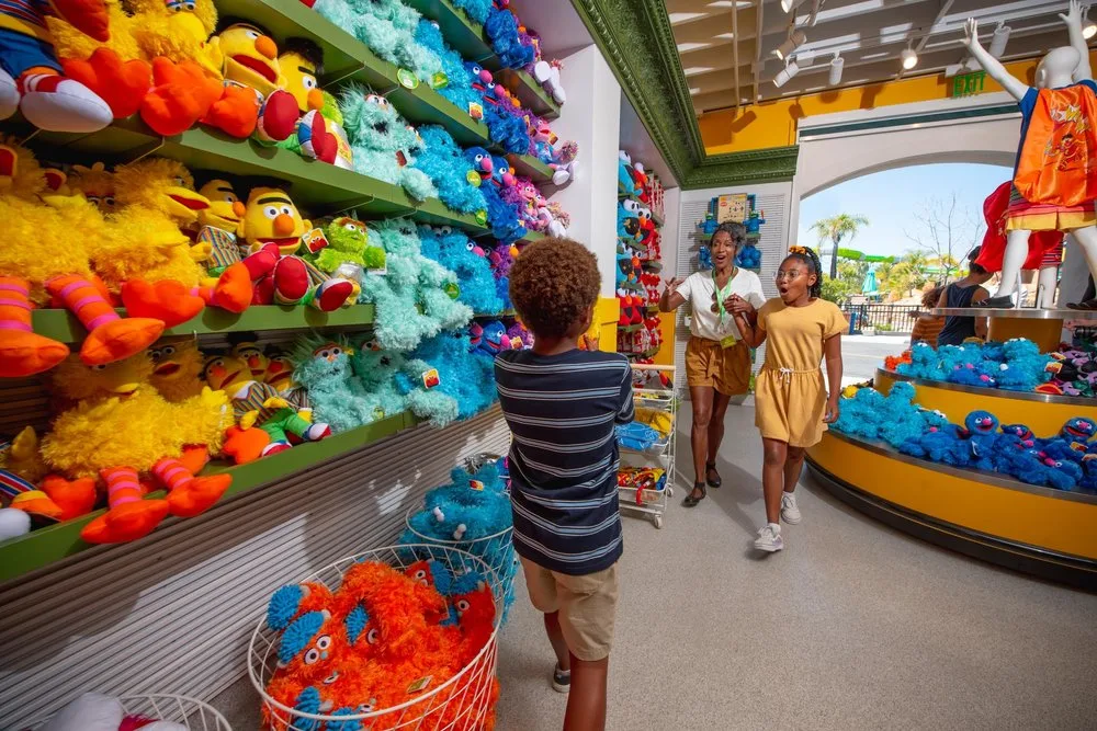 Children browsing a bright wall of Sesame Street plush toys in a theme park gift shop