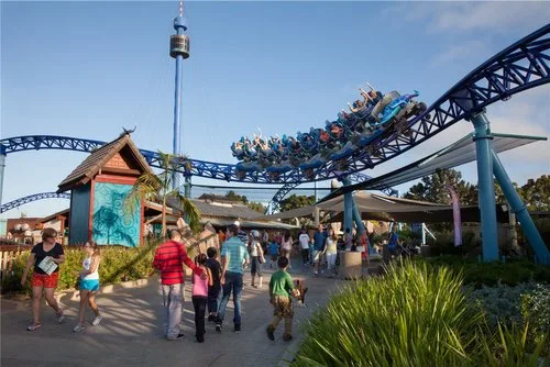 Guests walking along a landscaped path toward a blue roller coaster and themed entry building