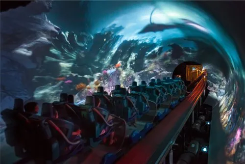 Riders in a dark ride vehicle beneath a curved projection screen of sea creatures