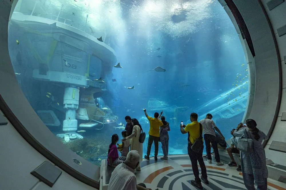 Guests photographing a vast aquarium tank with a submerged research station through a circular window