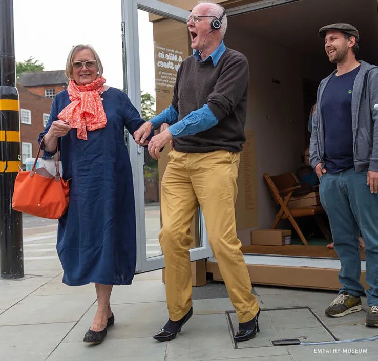 Visitors stepping out of the Empathy Museum's A Mile in My Shoes exhibit, one laughing in borrowed shoes and headphones