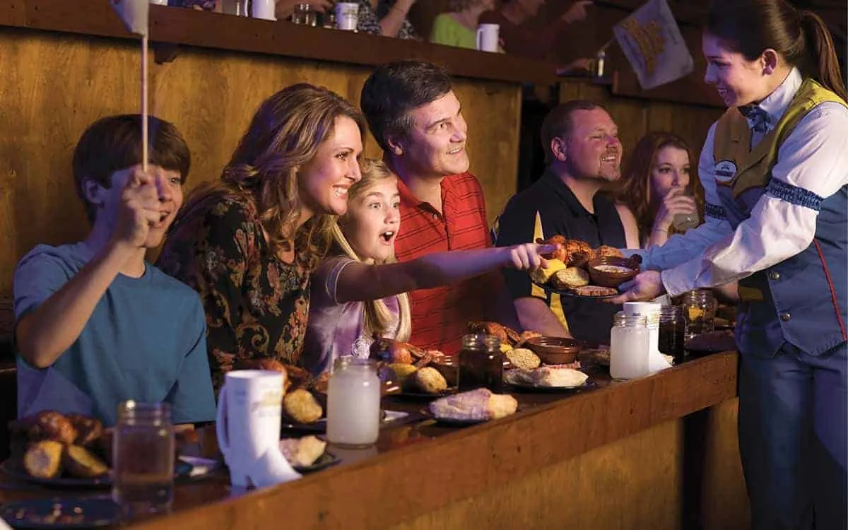 Family cheering at a dinner show as a costumed server passes food along a long wooden table