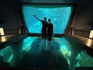 Two visitors posing on a glass floor in front of a large aquarium window at SeaWorld Abu Dhabi