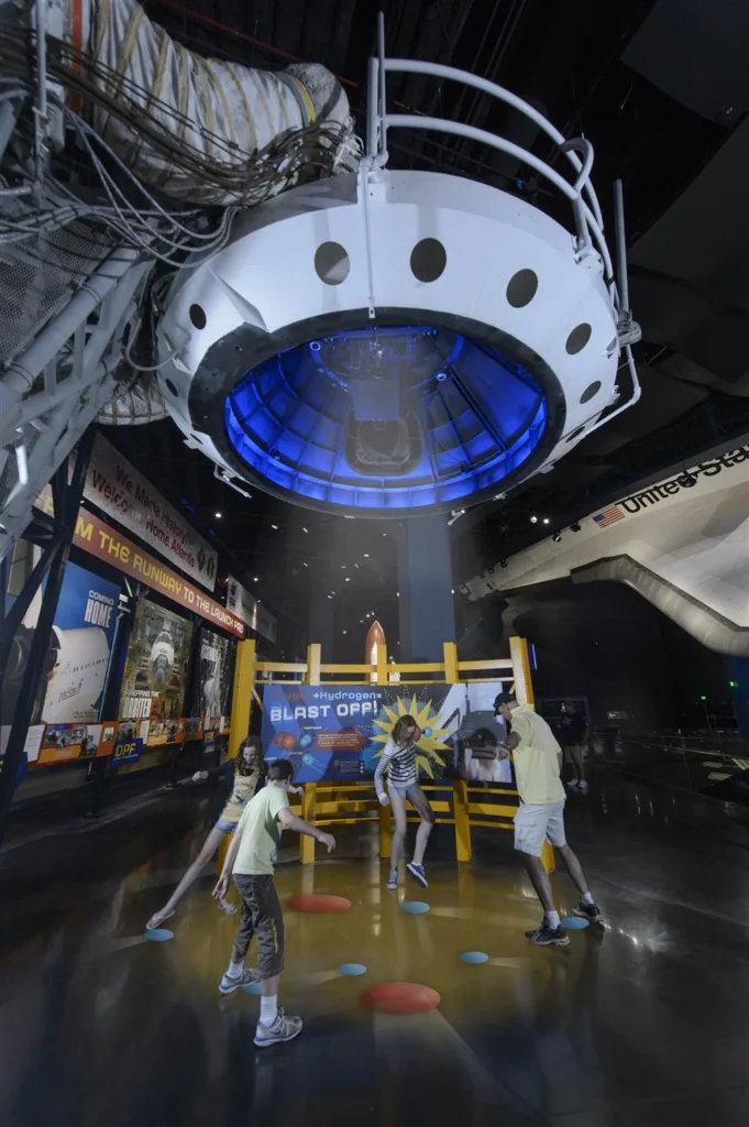 Children playing a floor game beneath a suspended spacecraft module in the Space Shuttle Atlantis exhibit