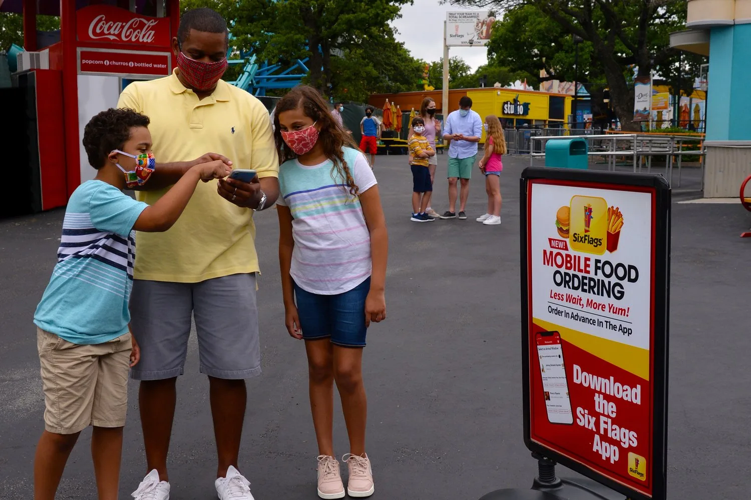 Masked father and kids ordering on a phone beside a Six Flags mobile food ordering sign
