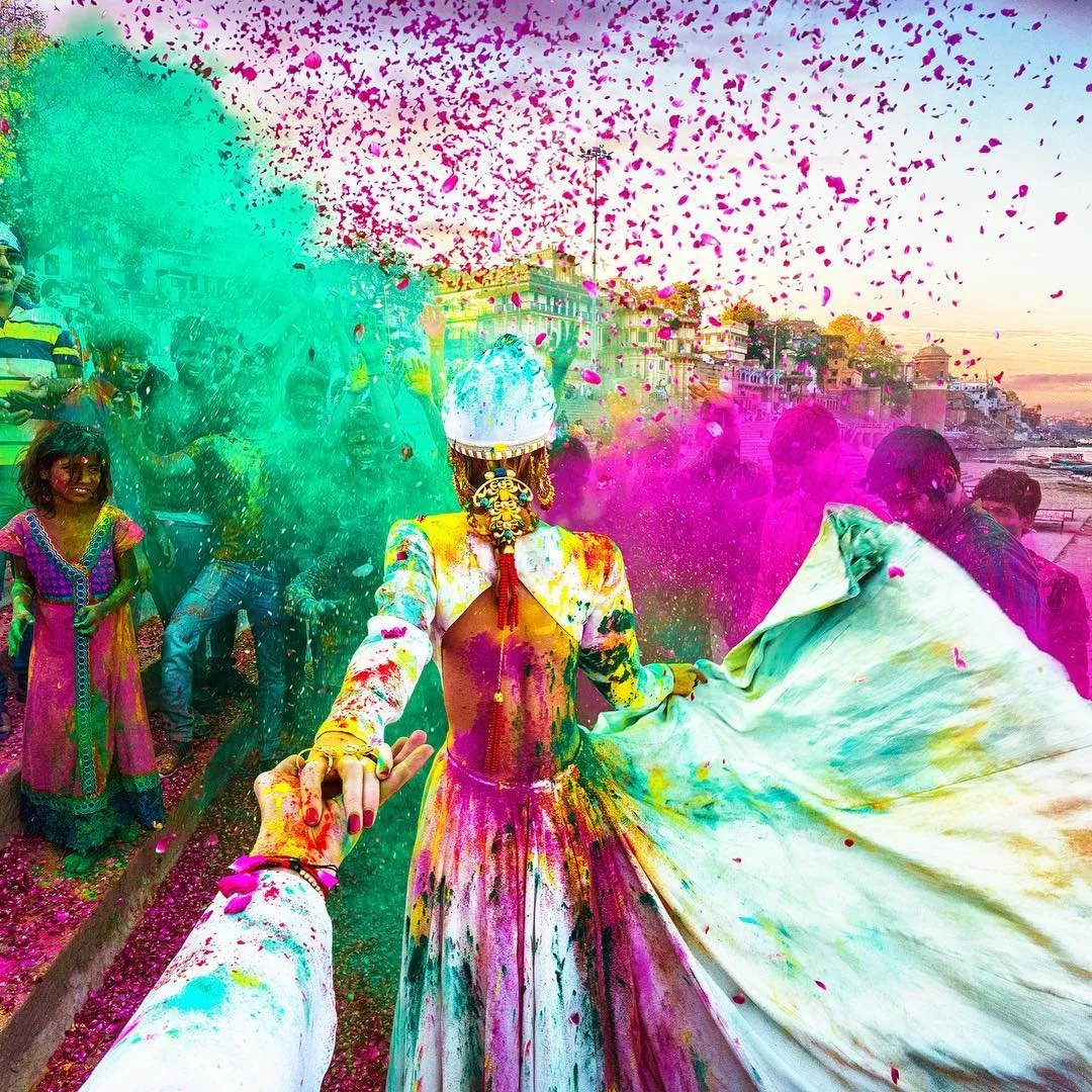 Follow-me style photo of a woman in a white dress leading the way through Holi color powder in Varanasi, India