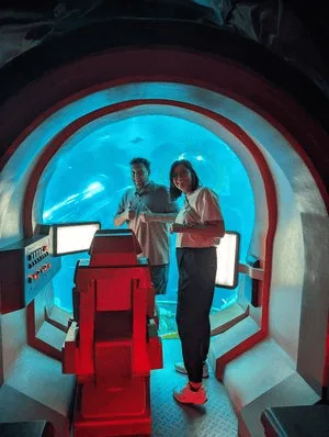 Two visitors standing in a red submarine-style cockpit before a large aquarium window at SeaWorld Abu Dhabi