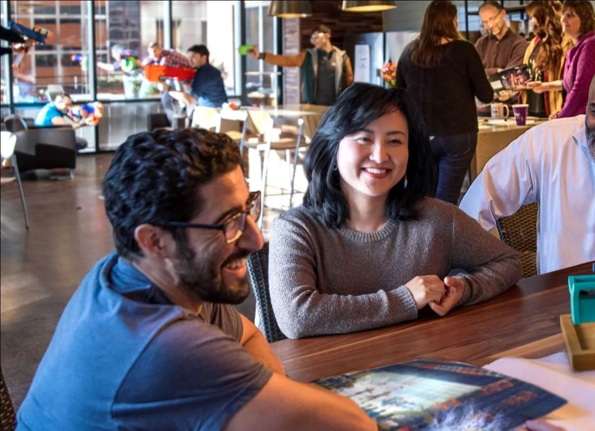 Smiling team members gathered around a table in a busy, sunlit office cafe