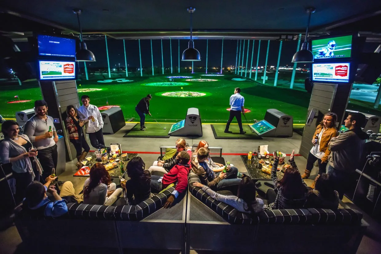 Friends lounging and golfers hitting balls in a bay overlooking a lit driving range at Topgolf