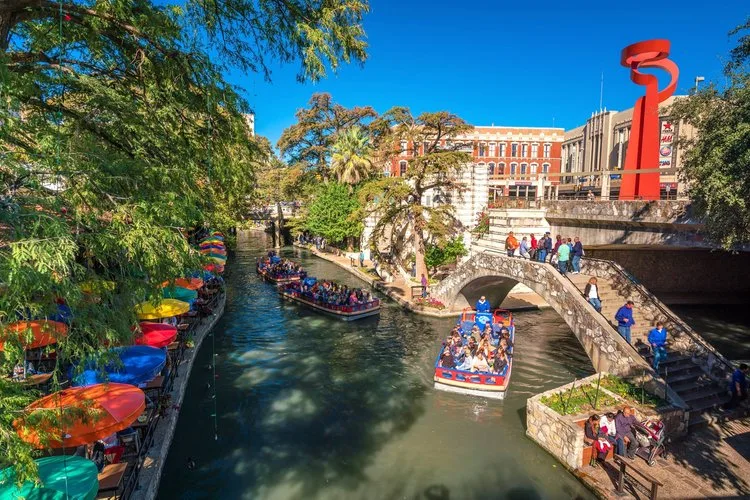 Tour boats on the San Antonio River Walk beneath cypress trees and the red Torch of Friendship sculpture