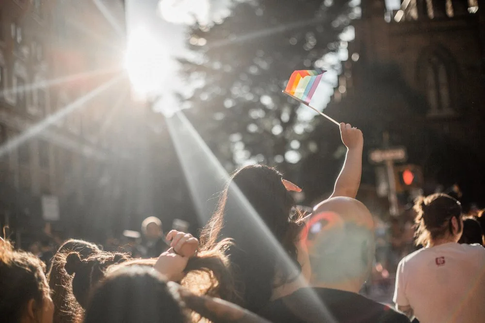 Crowd at a sunlit Pride event with a raised hand waving a small rainbow flag
