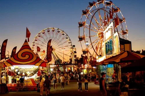 State fair midway with striped tents, food stands and lit Ferris wheels at dusk