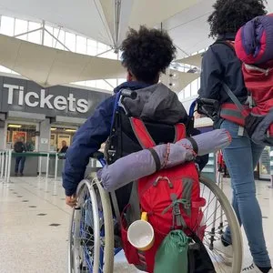 Wheelchair traveler with a loaded backpack heading toward airport ticket counters